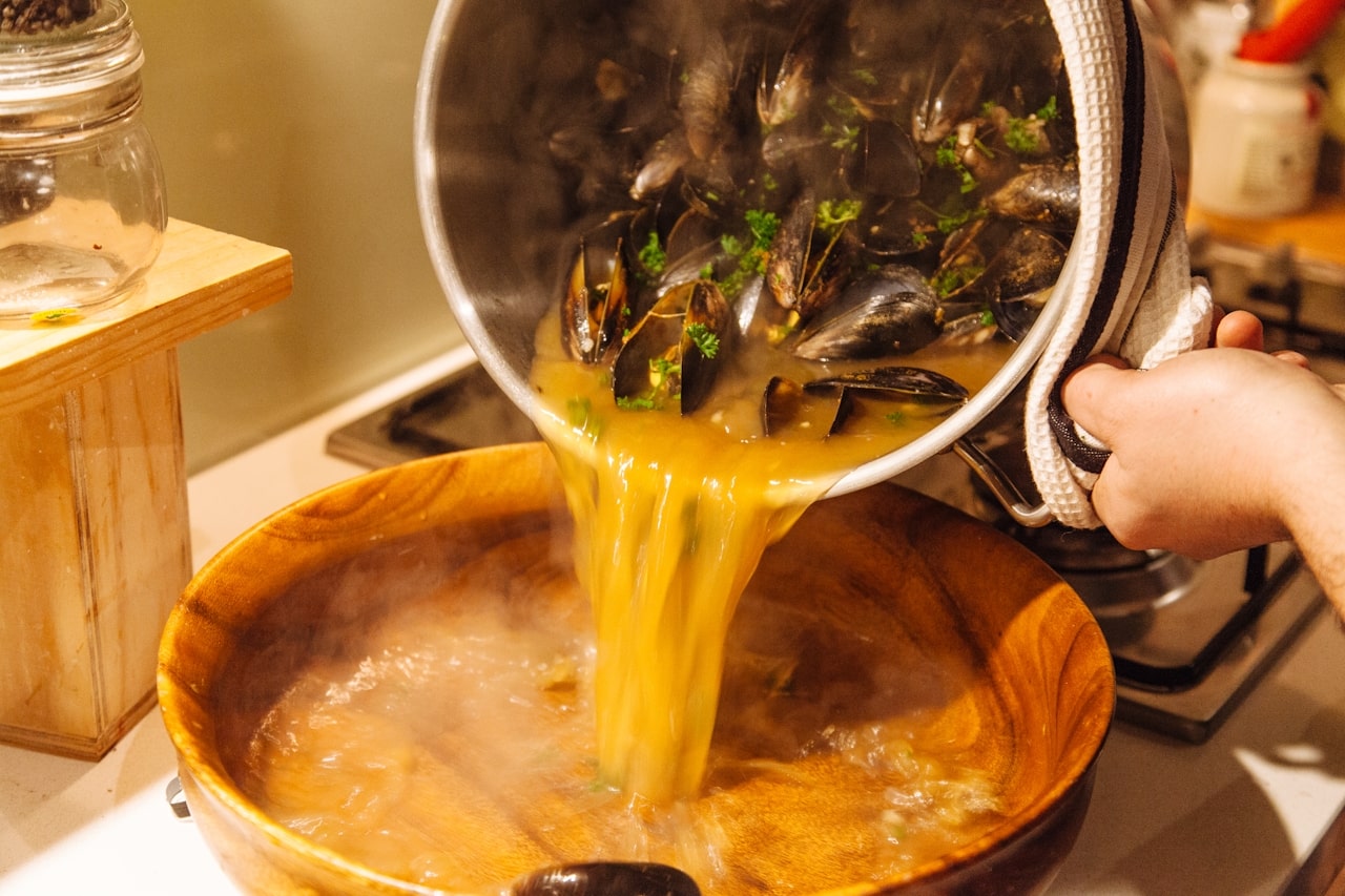 person pouring broth from pot of cooked mussels into wooden bowl