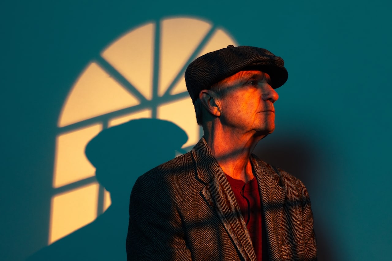 elderly man wearing herringbone blazer and messenger cap standing in front of turquoise wall