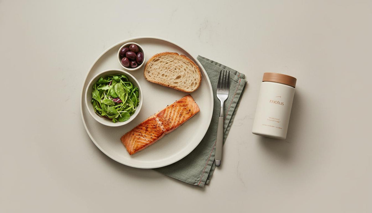 Overhead minimalist kitchen counter with Motus supplement beside grilled salmon, olives, mixed greens and whole-grain bread — how to improve liver metabolism