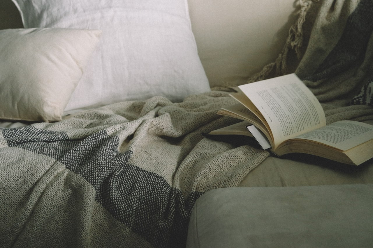 open paperback book lying on top of sofa with throw blanket and pillows