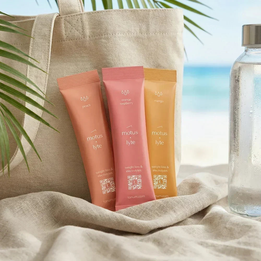 Three Tonum MOTUS LYTE electrolyte packets beside a water bottle and tote bag at the beach.